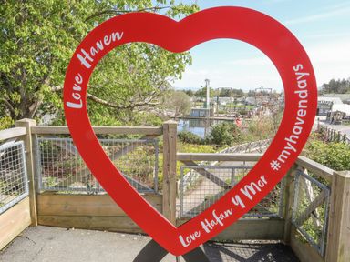A red heart-shaped frame with Love Haven and Love Hafan y Mor #myhavendays written on it with a park and water in the background