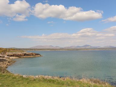 A coastal view with rocky shore and grassy foreground looking out over water towards distant hills and a partly cloudy sky at 171 Snowdonia View in Chwilog