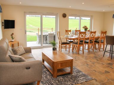 A living area with a sofa, coffee table, dining table with chairs, and sliding glass doors overlooking a green field at Holly Lodge in Forden near Montgomery