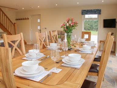 A dining room with a wooden table set for eight with white dishes and glasses and a vase of flowers at Holly Lodge in Forden near Montgomery