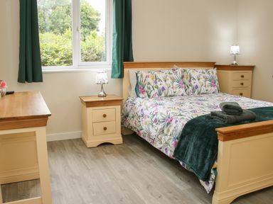 A bedroom with a bed with floral bedding two bedside tables with lamps a chest of drawers and a window with green curtains at Holly Lodge in Forden near Montgomery