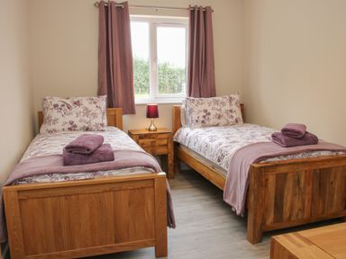 A bedroom with two single wooden beds with floral bedding purple towels and curtains and a wooden nightstand with a lamp at Holly Lodge in Forden near Montgomery