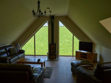 A living room with leather sofas a coffee table a wood stove and large triangular windows showing a grassy field at Holly Lodge in Forden near Montgomery