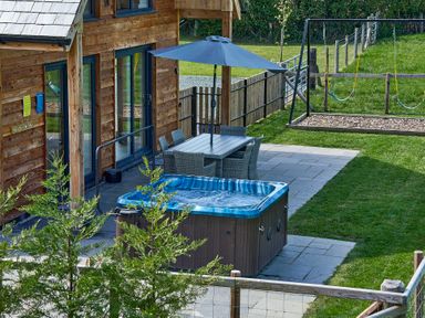 An outdoor patio with a hot tub a dining table with chairs and an umbrella next to a wooden house with a swing set in the grassy yard at Holly Lodge in Forden near Montgomery