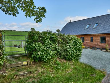 A wooden house with a slate roof next to a gravel driveway and a green hedge with a metal gate leading to a grassy field at Holly Lodge in Forden near Montgomery