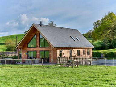 A wooden house with large triangular windows and a fenced yard with a swing in a rural setting at Holly Lodge in Forden near Montgomery