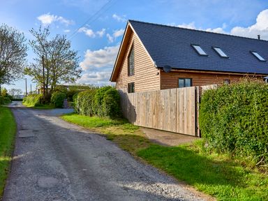 A wooden house with a slate roof next to a gravel road and hedges at Holly Lodge in Forden near Montgomery