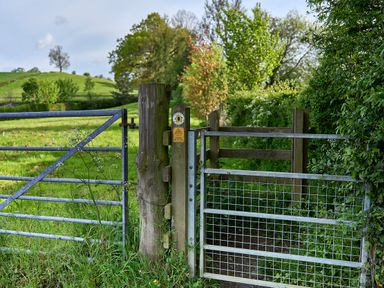 A metal gate and wooden fence posts with a countryside path and green fields in the background at Holly Lodge in Forden near Montgomery