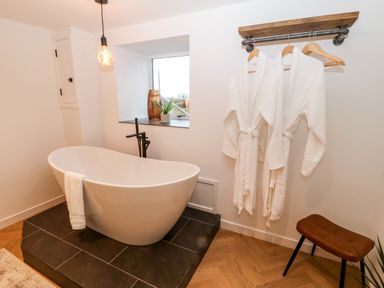 A bathroom with a freestanding bathtub on a tiled platform two white robes hanging on a wall rack a small window with a plant and vase and a brown stool at Sgubor in Llangefni