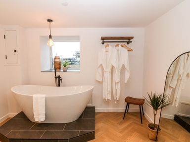 A bathroom with a freestanding bathtub a towel hanging on the tub two white bathrobes hanging on a wall rack a brown stool a tall mirror and a potted plant at Sgubor in Llangefni