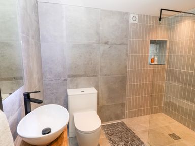 A bathroom with a white toilet and bowl sink next to a walk in shower with toiletries in a wall niche at Sgubor in Llangefni