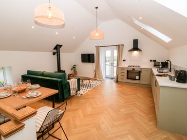 An open plan kitchen and living area with a dining table a green sofa a wall-mounted TV and kitchen cabinets at Sgubor in Llangefni