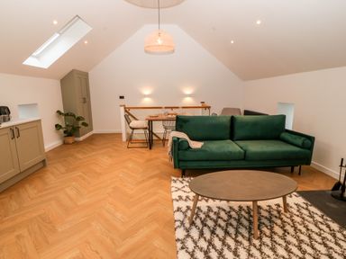 A living area with a green sofa and round coffee table on a patterned rug near a dining table with chairs and kitchen cabinets at Sgubor in Llangefni