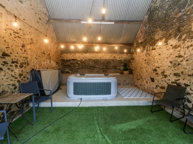A room with stone walls and a corrugated metal roof containing a hot tub on a raised platform with string lights overhead and chairs with a small table on artificial grass floor at Sgubor in Llangefni