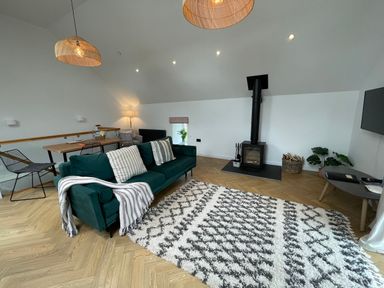 A living room with a green sofa and patterned rug near a wood stove and a dining table at Sgubor in Llangefni