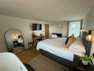 A bedroom with a bed, bedside tables, a large mirror, a wall-mounted TV, a desk with chair and stool, and a door at Sgubor in Llangefni