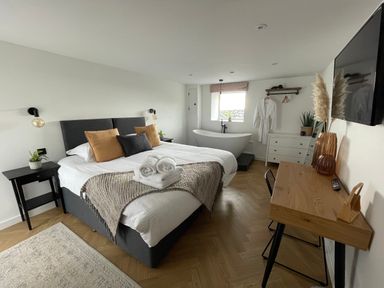 A bedroom with a bed and pillows a freestanding bathtub a desk with a chair and a wall-mounted television at Sgubor in Llangefni