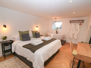 A bedroom with a double bed towels on the bed a bathtub near the window bathrobes hanging on the wall and a wooden desk with a chair at Beudy in Llangefni