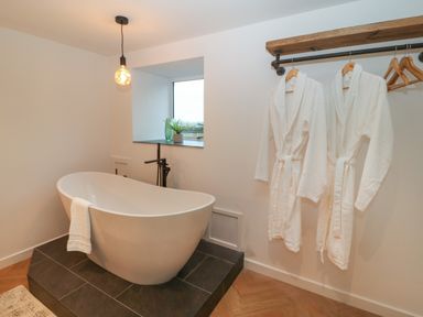 A bathroom with a freestanding bathtub on a tiled platform two bathrobes hanging on a wall rack and a small window with plants at Beudy in Llangefni