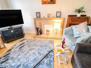 A living room with a television on a stand a tiled fireplace with decorations a blue armchair and a small table with snacks and wine at Grannys in Bushmills County Antrim