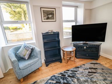 A corner of a living room with a blue armchair, a small wooden round table, a blue shelving unit, a flat screen TV on a blue TV stand, and two windows at Grannys in Bushmills County Antrim