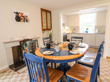 A dining area with a round table set with cups and plates surrounded by blue chairs next to a fireplace and an open doorway to a kitchen at Granny's in Bushmills County Antrim