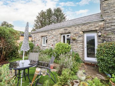 An outdoor garden area with a glass table umbrella and two chairs next to a stone wall house with plants at Cwm Caeth Cottage Nantmor near Beddgelert