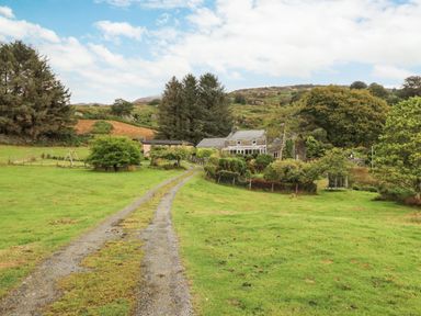 A gravel driveway leading to a stone house with a conservatory surrounded by trees and greenery at Cwm Caeth Cottage in Nantmor near Beddgelert