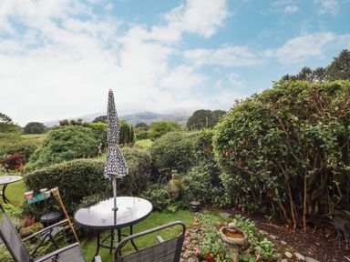 A garden with a round table and chairs and an umbrella surrounded by hedges and plants at Cwm Caeth Cottage in Nantmor near Beddgelert