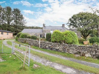 A stone cottage with a gravel driveway and stone wall surrounded by bushes and trees at Cwm Caeth Cottage in Nantmor near Beddgelert