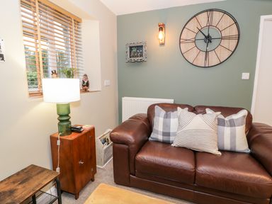 A living room with a brown leather sofa with pillows a green lamp on a wooden cabinet and a large wall clock at Cwm Caeth Cottage in Nantmor near Beddgelert