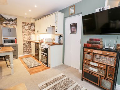 A kitchen area with white cabinets oven and microwave next to a decorative wooden cabinet with books and a wall mounted TV at Cwm Caeth Cottage in Nantmor near Beddgelert