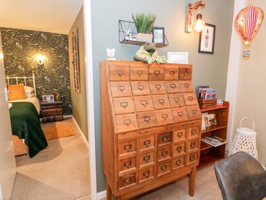A room with a wooden cabinet with many drawers a shelf with plants and toys and a view of a bedroom with a bed and patterned wallpaper at Cwm Caeth Cottage in Nantmor near Beddgelert