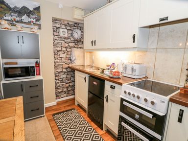 A kitchen with white cabinets a stove a microwave on a shelving unit and a stone patterned wall at Cwm Caeth Cottage Nantmor near Beddgelert