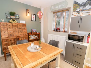 A dining area with a wooden table and chairs a cabinet with a microwave wooden drawers and shelves with books at Cwm Caeth Cottage in Nantmor near Beddgelert