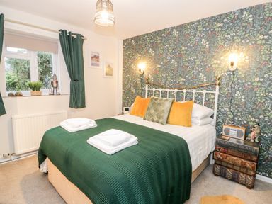 A bedroom with a double bed green bedspread green curtains floral wallpaper and a bedside table made of stacked books at Cwm Caeth Cottage Nantmor near Beddgelert