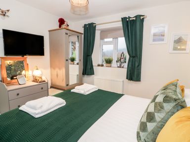 A bedroom with a bed covered with green and white bedding two folded towels on the bed a wardrobe with a mirror a dresser with a lamp and mirror a wall-mounted television and a window with green curtains at Cwm Caeth Cottage in Nantmor near Beddgelert