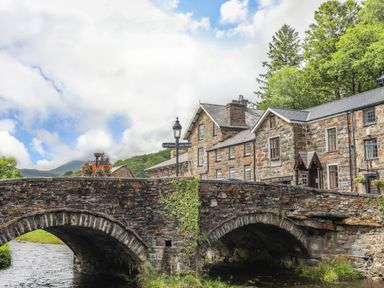 A stone bridge over a river with stone buildings and trees in the background at Cwm Caeth Cottage in Nantmor near Beddgelert