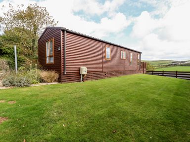 A wooden mobile home on a grass lawn with trees and fields in the background at Orchard View in Bossiney near Tintagel