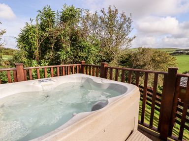 An outdoor hot tub on a wooden deck with wooden railing surrounded by trees and fields at Orchard View in Bossiney near Tintagel