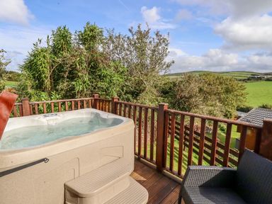 A hot tub on a wooden deck with chairs and green fields in the background at Orchard View in Bossiney near Tintagel