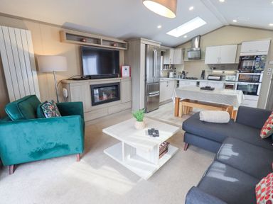 A living room and kitchen with a green armchair a black sofa a coffee table a TV above an electric fireplace and a dining table at Windermere View Pony Meadow 37 White Cross Bay near Windermere