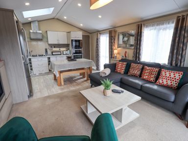 An open living room and kitchen area with a dark sectional sofa with patterned pillows a light wooden coffee table with a plant a dining table with a tablecloth and wooden benches and kitchen cabinets and appliances at Windermere View in Pony Meadow 37 White Cross Bay near Windermere