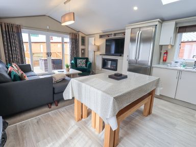 A kitchen and living area with a dining table covered with a cloth a grey couch a green armchair a television above a fireplace and a stainless steel fridge at Windermere View in Pony Meadow 37 White Cross Bay near Windermere