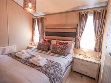 A bedroom with a bed with patterned pillows and folded towels on top and nightstands with lamps by windows with curtains at Windermere View in Pony Meadow 37 White Cross Bay near Windermere