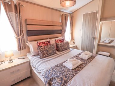 A bedroom with a bed with pillows and folded towels bedside tables with lamps curtains and a door at Windermere View in Pony Meadow 37 White Cross Bay near Windermere
