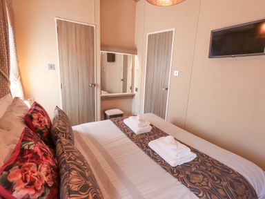 A bedroom with a bed with folded towels a wall mounted tv and two closed wooden doors at Windermere View in Pony Meadow 37 White Cross Bay near Windermere