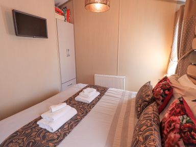 A bedroom with a double bed, folded towels, a wardrobe, a wall-mounted television and a window with curtains at Windermere View in Pony Meadow 37, White Cross Bay near Windermere
