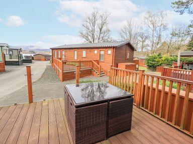 An outdoor wooden deck with brown wicker table and railing multiple wooden cabins and trees in the background at Windermere View Pony Meadow 37 White Cross Bay near Windermere