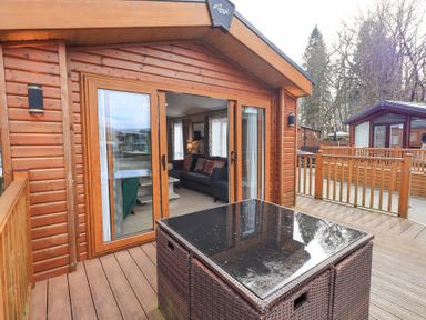An outdoor wooden deck with a dark wicker table in front of sliding glass doors leading to a living area at Windermere View in Pony Meadow 37 White Cross Bay near Windermere